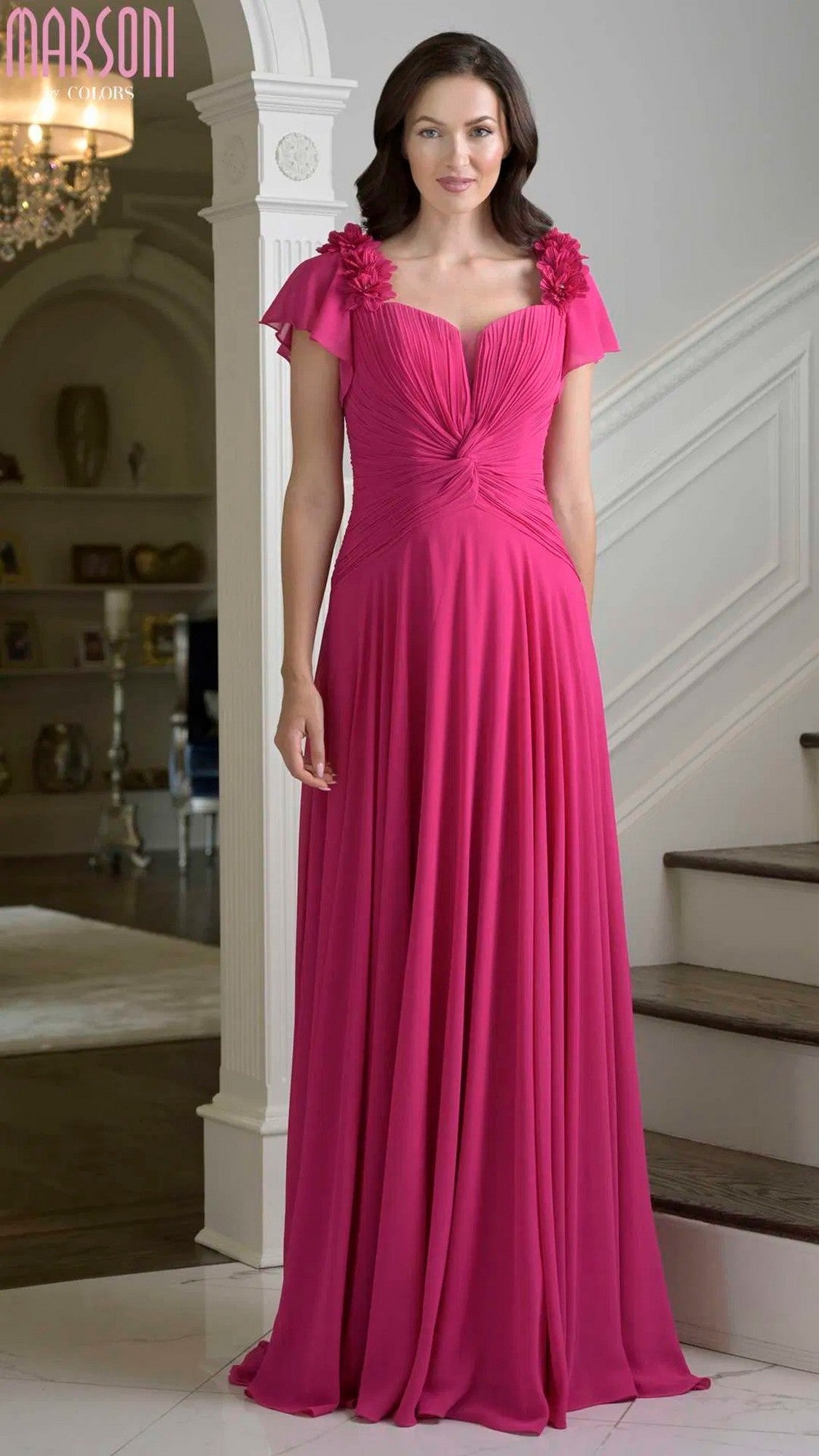 A woman stands indoors wearing the Marsoni M338 long formal dress in fuchsia, featuring short sleeves, an a-line chiffon skirt, and bright pink 3D floral appliques on the shoulders.